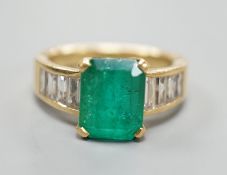A modern yellow metal and single stone emerald set dress ring, with graduated baguette cut diamond