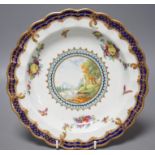 A Worcester plate painted with a landscape surrounded by fruit and moths under a blue and gilt