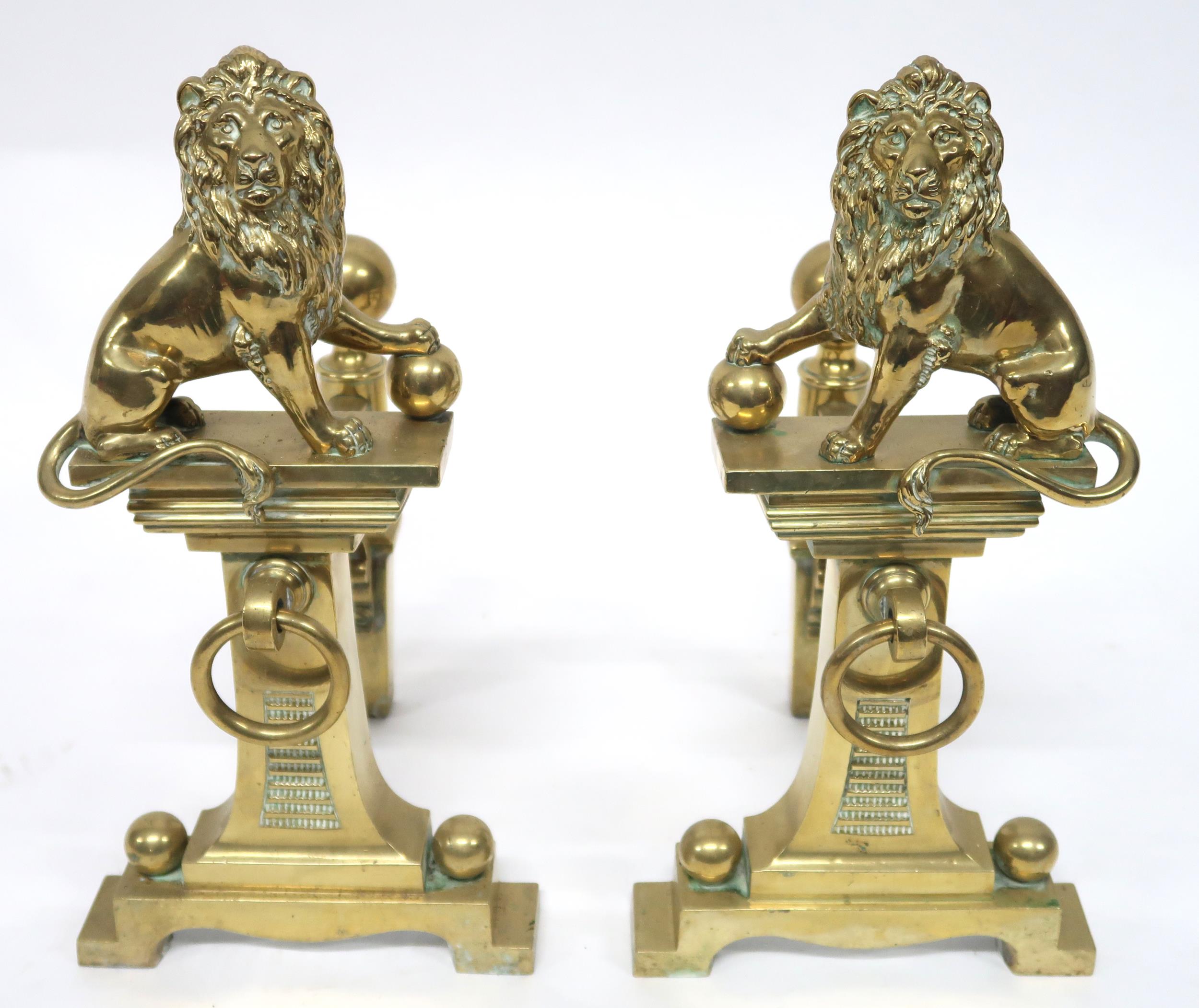A PAIR OF EARLY 20TH CENTURY CAST BRASS FIRE DOGS mounted with figural lions, 46cm high x 23cm