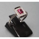AN ART DECO PLAQUE RING, the central princess cut ruby open back collet set to a border of sixteen