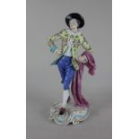 A Chelsea style porcelain figure of a youth wearing a yellow floral jacket and carrying a cape, gold