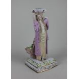 A Continental porcelain candlestick in the form of a woman with arms raised before a pillar, with