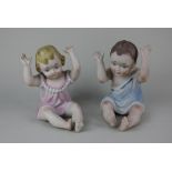 A pair of bisque figures of children, seated with raised arms 17cm high