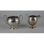 A George V silver sugar bowl and matching cream jug circular pedestal form engraved with festoons of