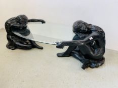 A DESIGNER COFFEE TABLE, THE OVAL GLASS TOP SUPPORTED BY TWO KNEELING NUDE FEMALE FIGURES.