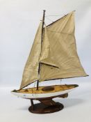 A WOODEN MODEL OF A YACHT WITH CANVAS SAILS ON DISPLAY STAND LENGTH 60CM. HEIGHT 82CM.