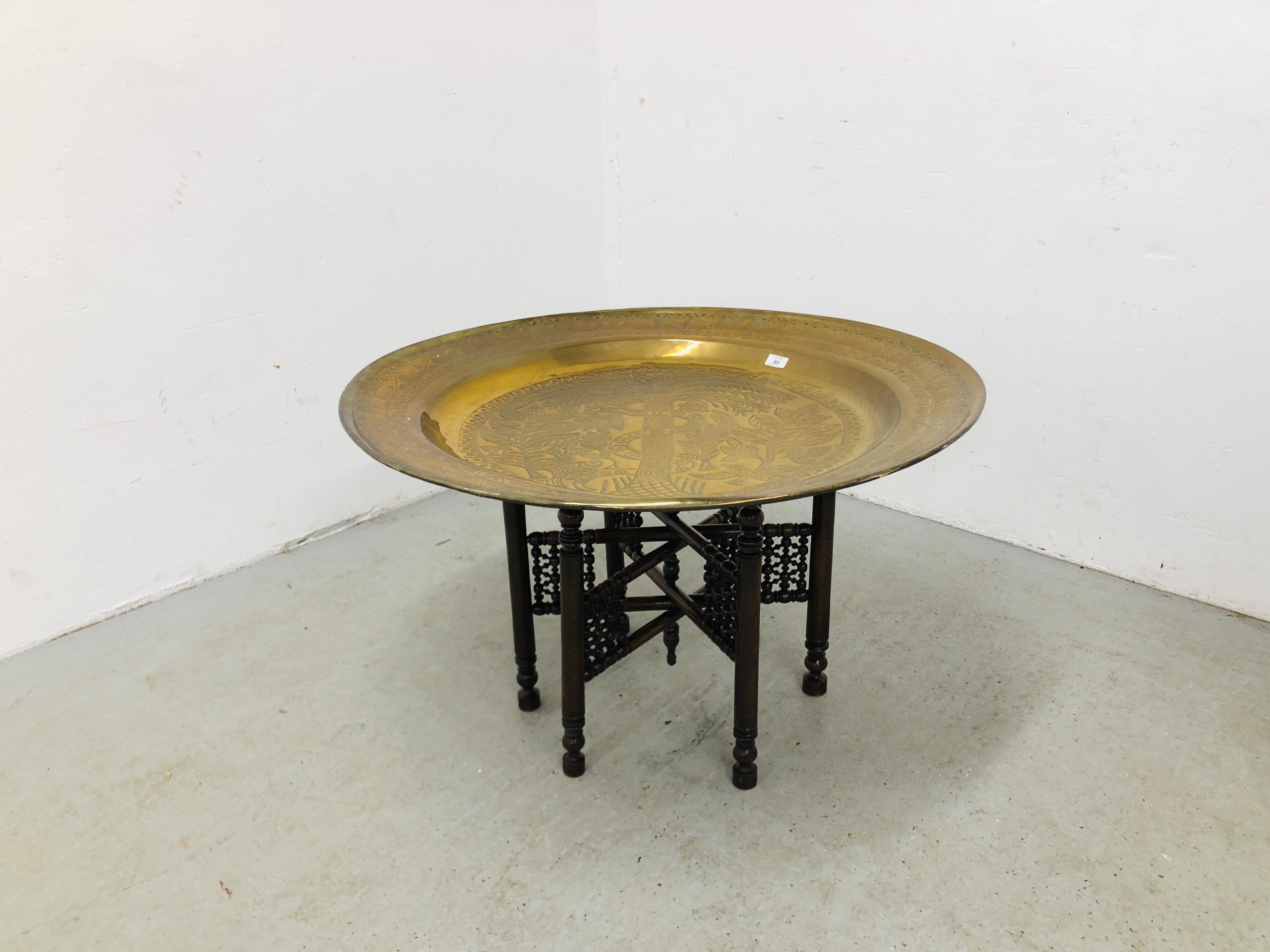 A LARGE BRASS EASTERN TRAY TABLE ON FOLDING CARVED WOODEN BASE (TRAY DIA. 91CM.