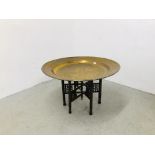 A LARGE BRASS EASTERN TRAY TABLE ON FOLDING CARVED WOODEN BASE (TRAY DIA. 91CM.