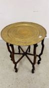 AN ORIENTAL BRASS CIRCULAR TABLE ON FOLDING CARVED ELM BASE, PROBABLY CHINESE, DIAMETER 56CM.