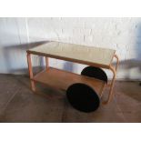 A Finmar Ltd Alvar Allto birch trolley with glass to top, black wheels and handle and label