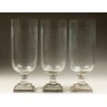 A SET OF THREE CUT GLASS STORM LAMPS on a square stepped bases. 15ins high.