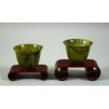 A PAIR OF CHINESE SPINACH JADE CUPS, with two wooden stands, cups 6cm diameter.
