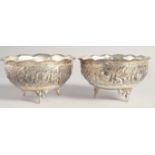 A PAIR OF WHITE METAL CIRCULAR BOWLS, with wavy rims, repousse decoration on three curving legs,