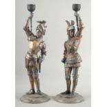 A PAIR OF PAINTED AND GILDED CANDLESTICKS men in suits of armour on circular bases. 16ins high.