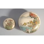 TWO JAPANESE SATSUMA CIRCULAR BOXES, the larger painted with flora over water with mountains beyond,