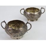 A pair of Indo-Asian white metal twin handled salts having village pastoral scenes upon with palm