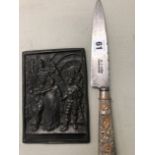 AN ANTIQUE WHITE METAL CHASED HANDLE ARGENTINIAN KNIFE TOGETHER WITH A BRONZE RELIEF CASTED PLAQUE.