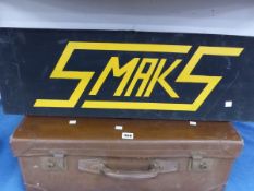 A BLACK GROUND WOODEN SIGN FOR SMAKS RAISED IN YELLOW TOGETHER WITH A SUITCASE