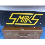 A BLACK GROUND WOODEN SIGN FOR SMAKS RAISED IN YELLOW TOGETHER WITH A SUITCASE