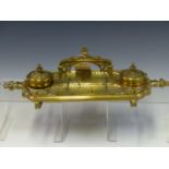 A GOTHIC REVIVAL BRASS INKSTAND WITH RECEIVERS FLANKING THE CENTRAL HANDLE RAISED ABOVE PEN TRAYS. W