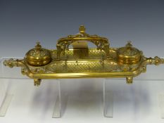 A GOTHIC REVIVAL BRASS INKSTAND WITH RECEIVERS FLANKING THE CENTRAL HANDLE RAISED ABOVE PEN TRAYS. W