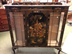 A 1920S BLACK LACQUER GROUND CHINOISERIE THREE DOOR DISPLAY CABINET DECORATED WITH ISLAND SCENES,