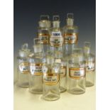 NINE CYLINDRICAL CLEAR GLASS PHARMACY BOTTLES AND STOPPERS, EACH WITH A RECESSED LABEL IN BLACK