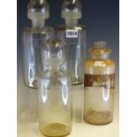 THREE CLEAR GLASS PHARMACY BOTTLES WITH BULBOUS CUP STOPPERS, THE CYLINDRICAL BODIES ENGRAVED OL: