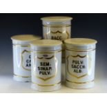 FOUR WHITE PORCELAIN CYLINDRICAL PHARMACY JARS AND COVERS LABELLED IN BLACK WITHIN GILT SHIELDS: