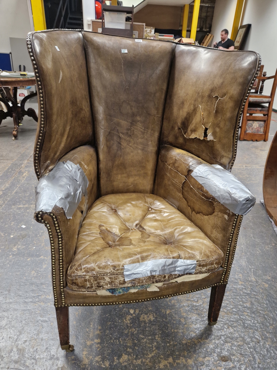 AN ANTIQUE LEATHER UPHOLSTERED WING ARMCHAIR WITH THE BACK ROUNDING OVER THE ARMS, THE SQUARE