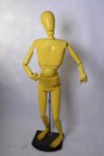 Vintage yellow painted wooden crash test dummy with multi-jointed body set on a metal stand, 179cm