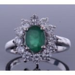 Emerald and diamond cluster ring, the oval faceted cut emerald within a small brilliant cut