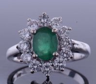 Emerald and diamond cluster ring, the oval faceted cut emerald within a small brilliant cut