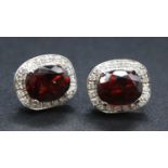 A pair of white metal, garnet and diamond oval cluster earrings, each featuring an oval faceted