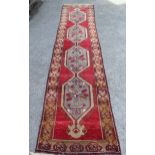 Belouch runner with a hooked pole medallion design on a red ground with borders, 12ft x 2ft 8ins