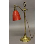 AN ADJUSTABLE BRASS STUDENTS LAMP, with a red metal shade on a circular base, height 55cm