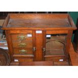 AN OAK SMOKERS CABINET COMPRISING TWO GLAZED CUPBOARD DOORS, THE LEFT OPENING TO FOUR DRAWERS, THE