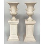 URNS ON STANDS, a pair, campana form stone effect on plinths, 46cm W x 135cm H.