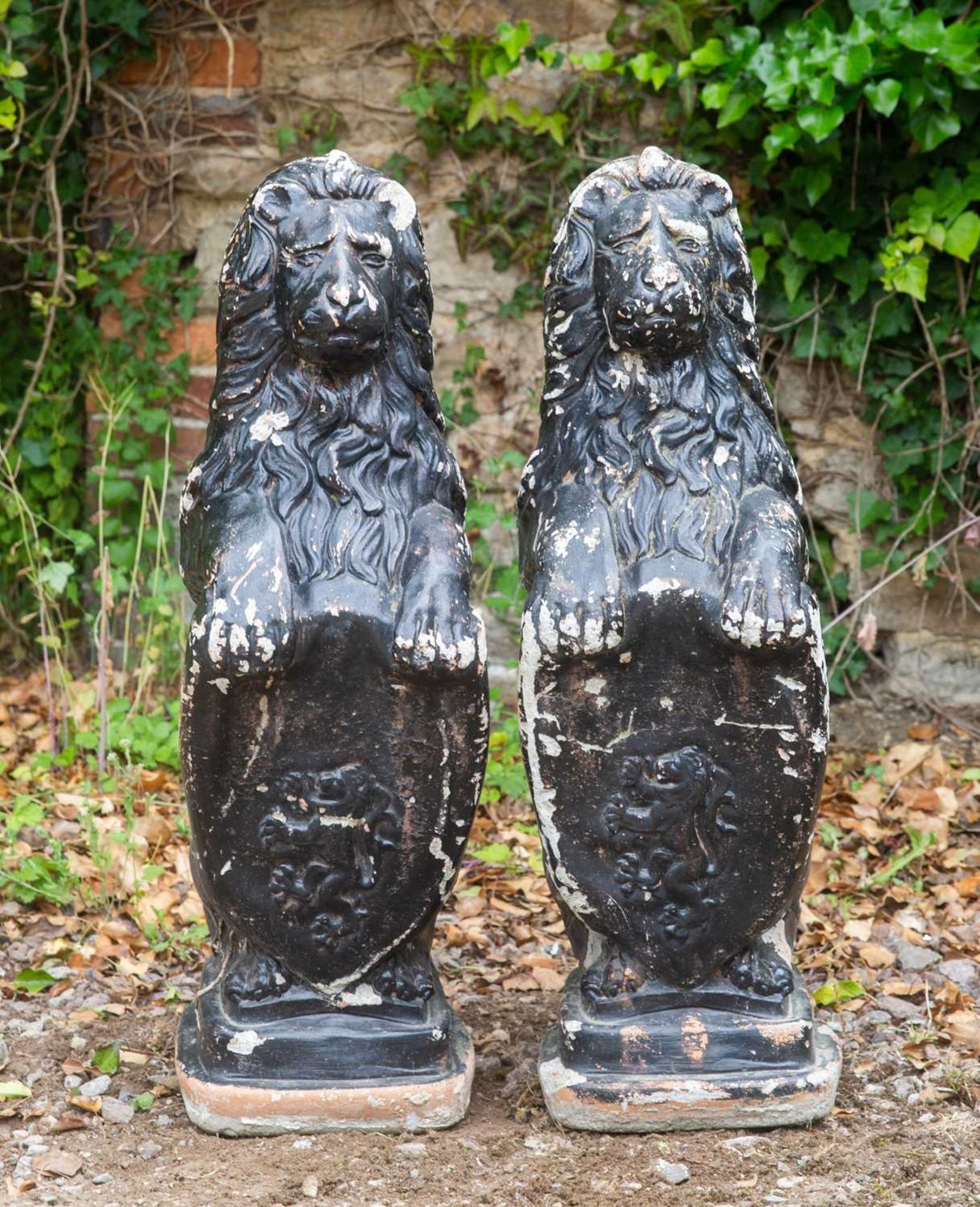 A pair of black painted cast reconstituted stone heraldic lions seated holding shields, each