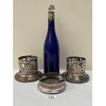 A pair of plated bottle coasters, another coaster and a blue glass wine bottle with white metal vine