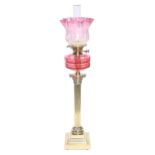 An Edwardian brass Corinthian column oil lamp, with cranberry glass fount, brass burner and an