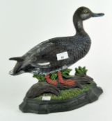 A painted cast iron duck door stop, the duck standing on a tree trunk on a grassy bank