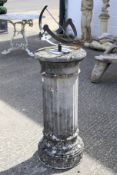 A grey metal sun dial with engraved Roman numerals. Fixed to a composite stone pedestal base.