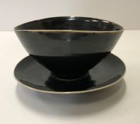 A Lucie Rie black glazed elliptical bowl and saucer, both bearing Lucie Rie marks, the bowl 14.