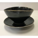 A Lucie Rie black glazed elliptical bowl and saucer, both bearing Lucie Rie marks, the bowl 14.