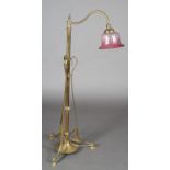 AN ART NOUVEAU BRASS STANDARD LAMP having a scrolled arm with cranberry glass shade, on a telescopic