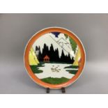 A pottery plaque in the style of Clarice Cliff depicting a lake and mountain landscape within an