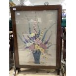 A 1930's oak fire screen table inset with a needlework panel of summer flowers held in a blue