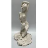 A white composite figure of a semi-naked art deco maiden, one arm raised over her head and poised on