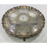 A circular silver tray with shaped rim, foliate engraving and central ‘R’, raised on four pad